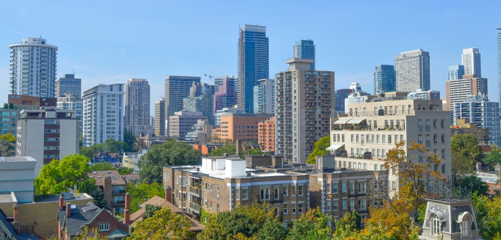 Cityscape view featuring various modern high-rise buildings and residential structures with trees interspersed throughout the area under a clear blue sky.