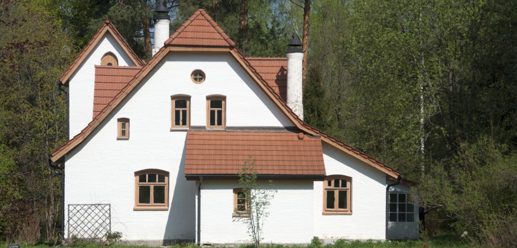 A charming white two-story house with brown-shingled roofs, tucked gracefully among trees and greenery, almost like a scene from Le Pas de Deux. The home features several picturesque windows and two stately chimneys.